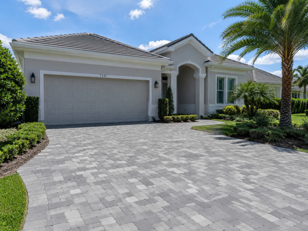 Gray tumbled paver driveway curving up to the two car garage of a home framed by clipped hedges and palms in Pompano Beach
