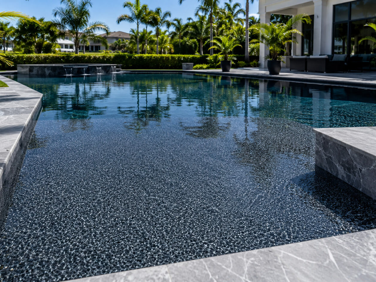 Dark charcoal pebble pool finish under still water, framed by gray marble coping and spillway on a Boca Raton property