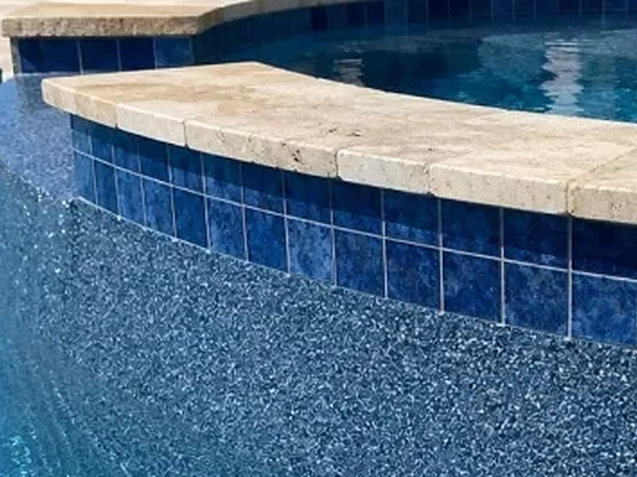 Blue pebble pool finish meeting a raised spa wall of deep blue tile capped with travertine coping for a Pompano Beach homeowner