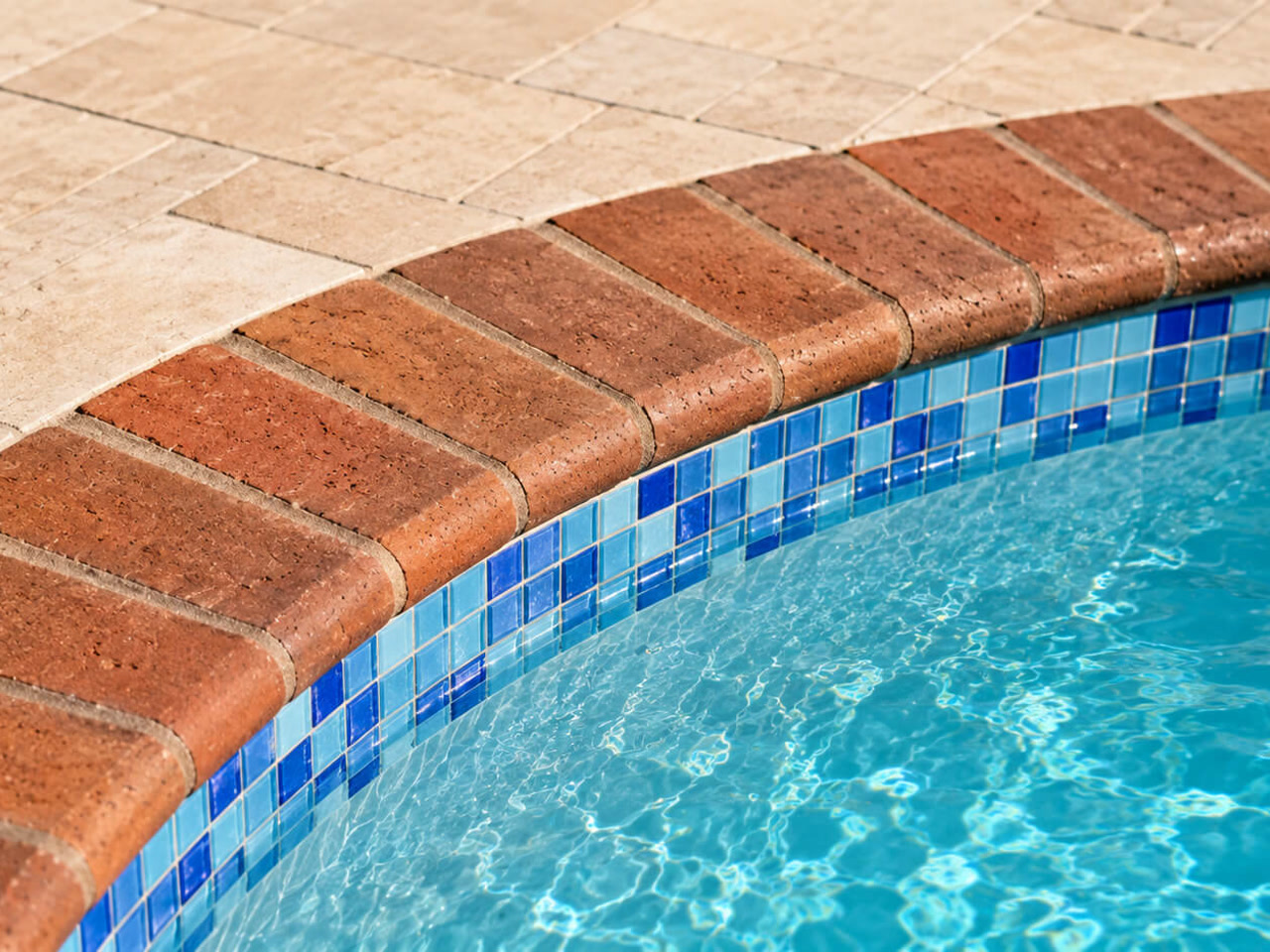Red brick coping following the curve of a pool over a blue mosaic waterline tile, beside a travertine deck at a Boca Raton home