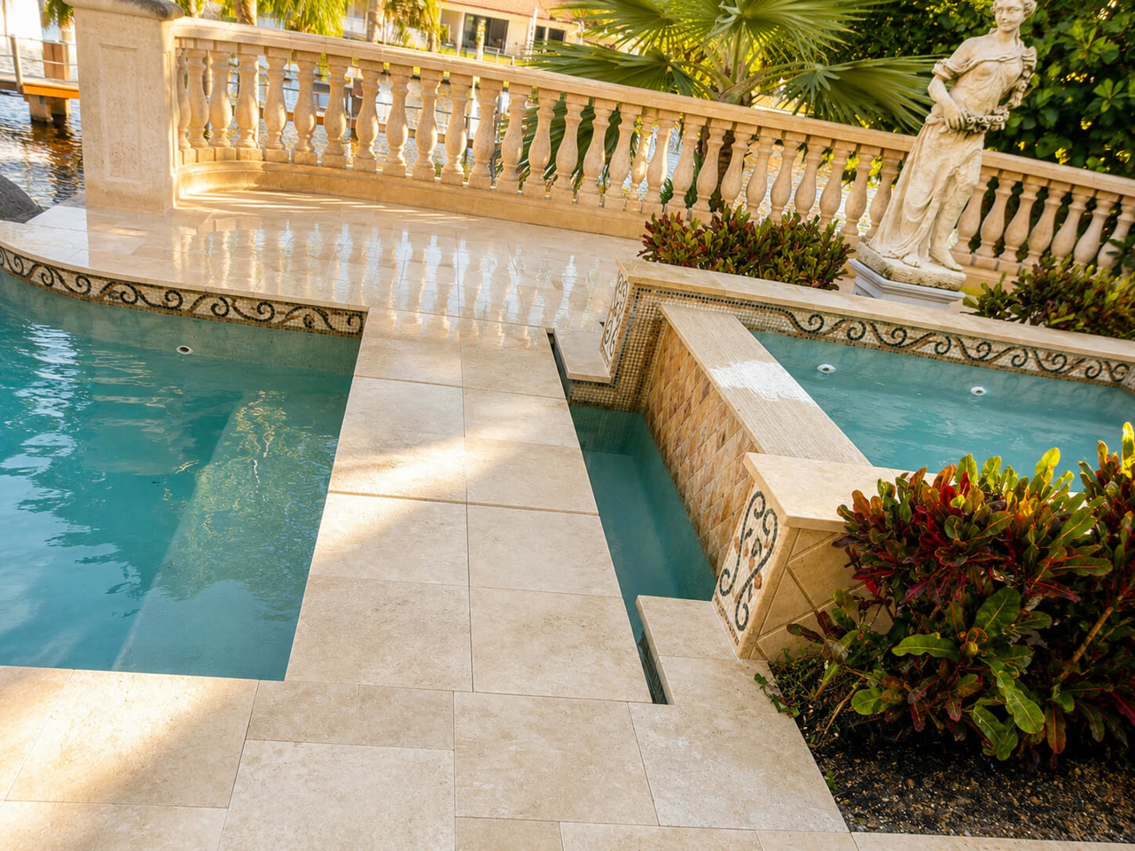 Ivory paver pool deck with a stone balustrade and mosaic tile spillway at a waterfront estate