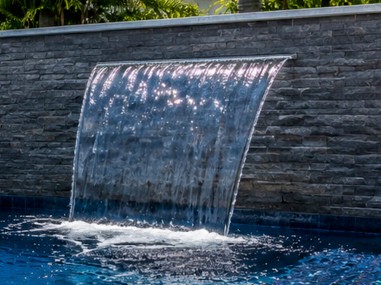 Sheer descent waterfall blade falling from a gray stacked stone wall into a deep blue pool